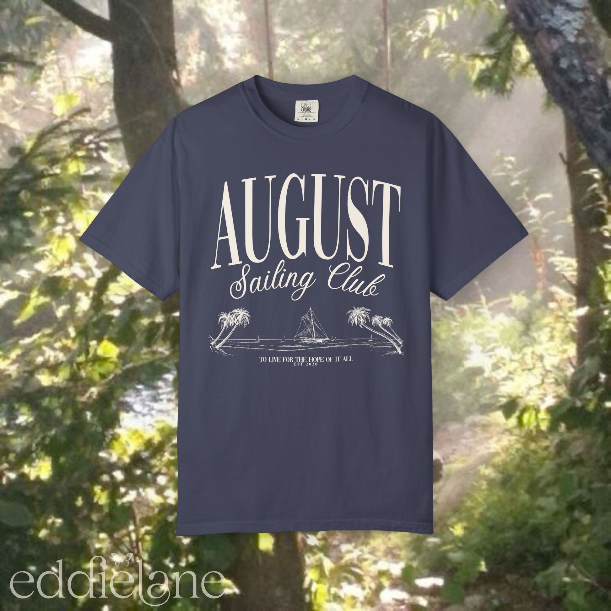 A minimalistic navy graphic tee that says 'August Sailing Club' with a sketch design of sailboats on a beach. Inspired by August from Folklore by Taylor Swift.