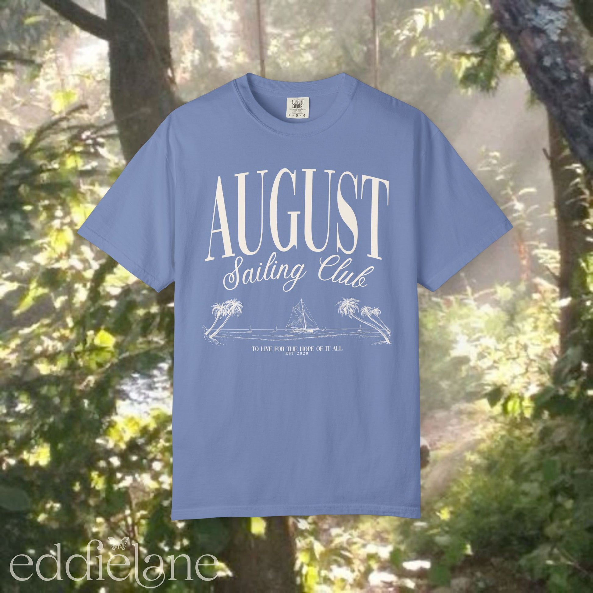 A minimalistic blue graphic tee that says 'August Sailing Club' with a sketch design of sailboats on a beach. Inspired by August from Folklore by Taylor Swift.