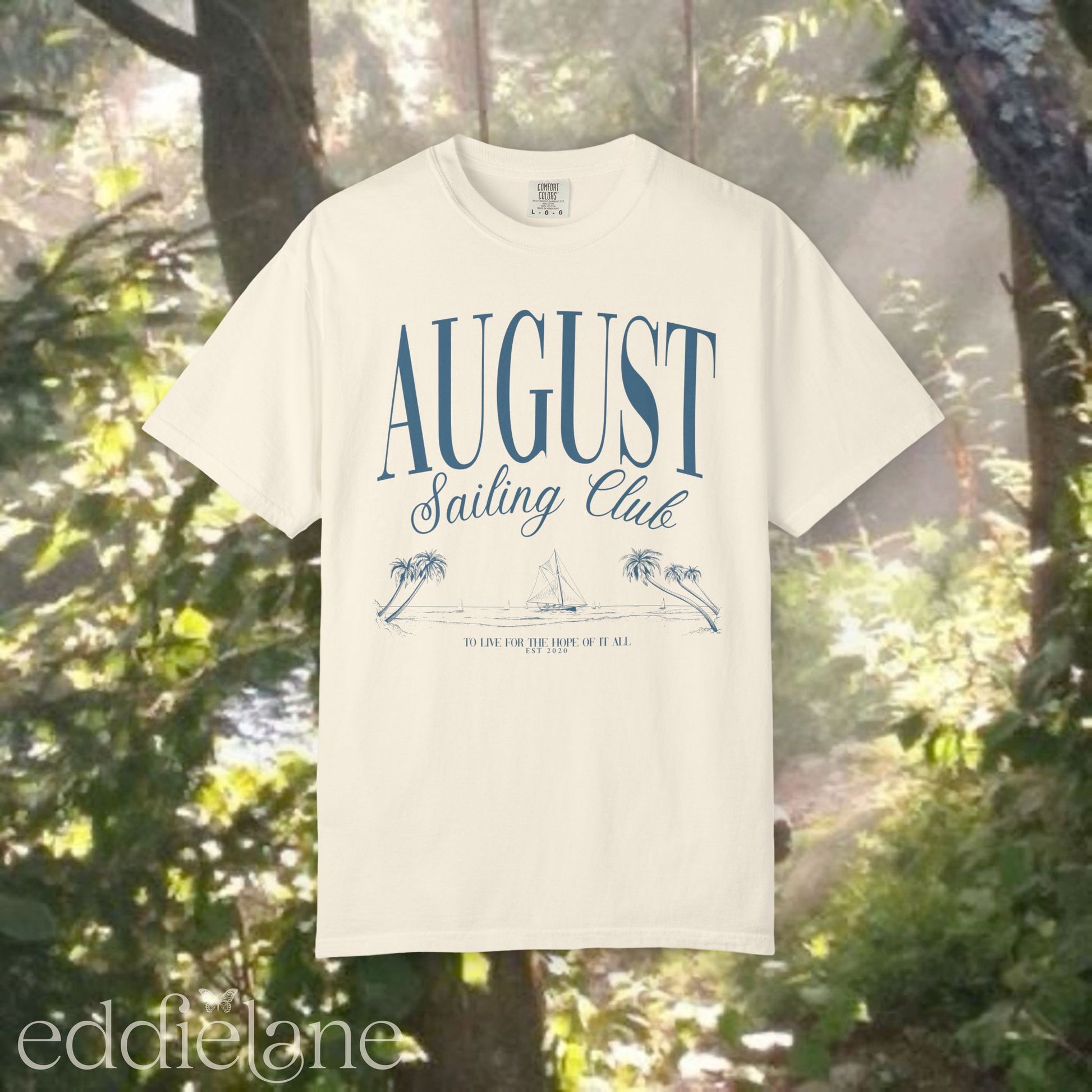 A minimalistic white graphic tee that says 'August Sailing Club' with a sketch design of sailboats on a beach. Inspired by August from Folklore by Taylor Swift.