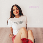 A woman sitting on a light wood floor against a white wall, wearing a light gray graphic T-shirt that reads 'HATING TAYLOR SWIFT DOESN'T MAKE YOU DEEP' and red-and-white gingham shorts with red cowboy boots. The brand name 'eddielane' appears in the upper right.