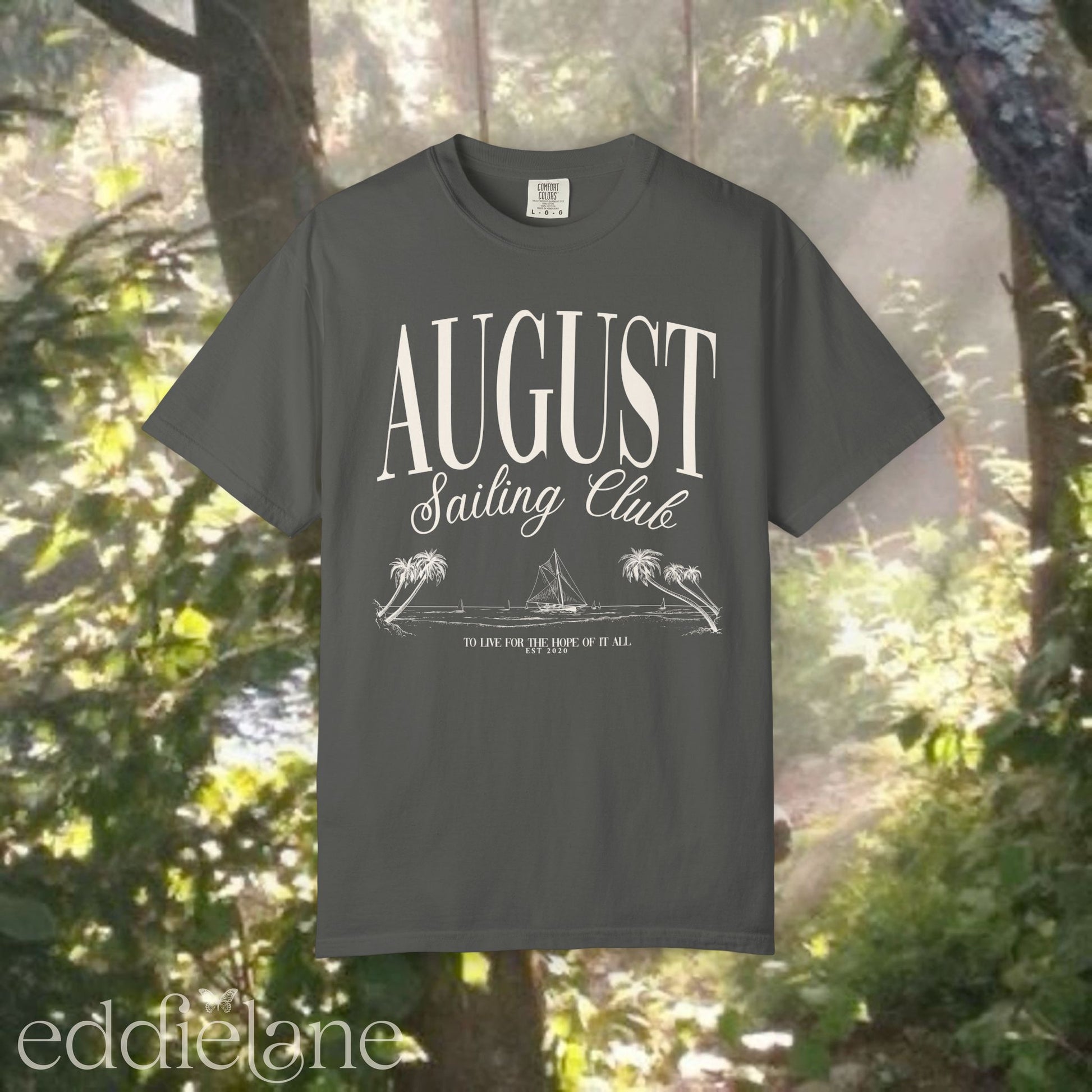 A minimalistic gray graphic tee that says 'August Sailing Club' with a sketch design of sailboats on a beach. Inspired by August from Folklore by Taylor Swift.