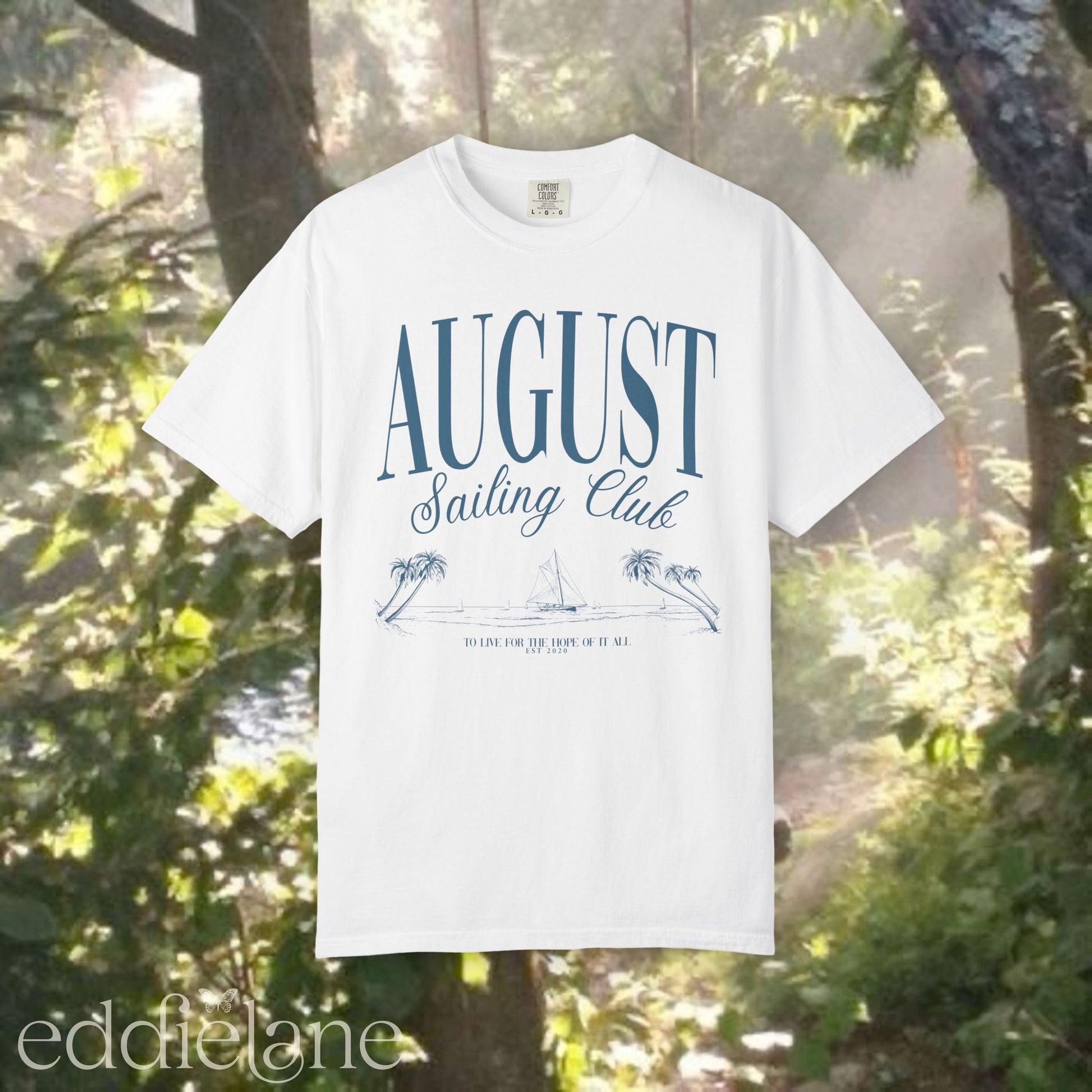 A minimalistic white graphic tee that says 'August Sailing Club' with a sketch design of sailboats on a beach. Inspired by August from Folklore by Taylor Swift.