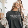 A woman wearing a dark charcoal gray graphic t-shirt with the text 'HATING TAYLOR SWIFT DOESN'T MAKE YOU DEEP' in white. She is wearing round gold-framed sunglasses, a cream knit cardigan, and holding a glass of iced coffee.