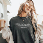 A woman wearing a dark charcoal gray graphic t-shirt with the text 'HATING TAYLOR SWIFT DOESN'T MAKE YOU DEEP' in white. She is wearing round gold-framed sunglasses, a cream knit cardigan, and holding a glass of iced coffee.
