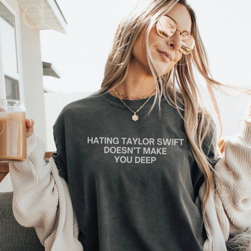A woman wearing a dark charcoal gray graphic t-shirt with the text 'HATING TAYLOR SWIFT DOESN'T MAKE YOU DEEP' in white. She is wearing round gold-framed sunglasses, a cream knit cardigan, and holding a glass of iced coffee.