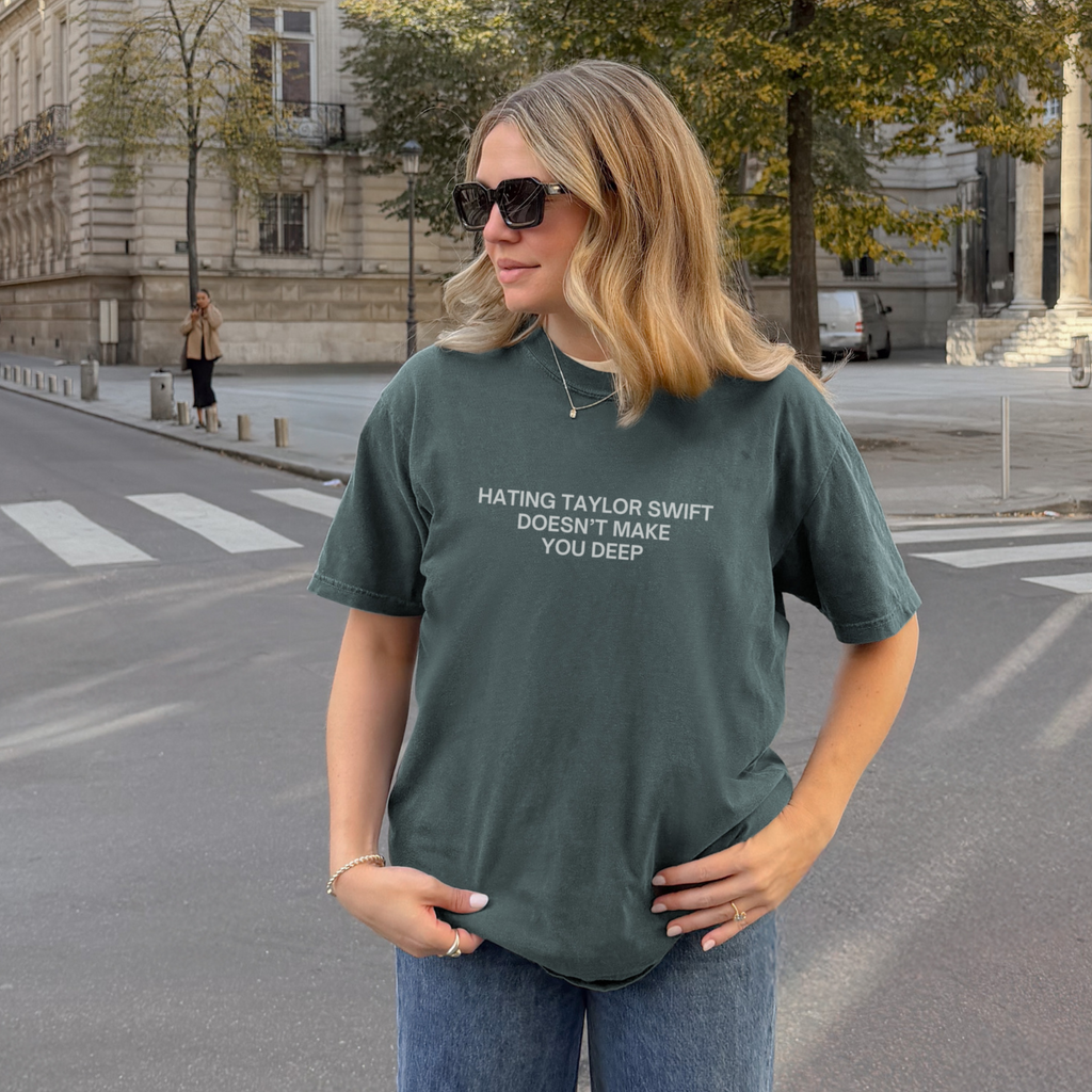 A woman wearing a dark green graphic t-shirt with the text 'HATING TAYLOR SWIFT DOESN'T MAKE YOU DEEP' stands on a city street in front of a historic building.