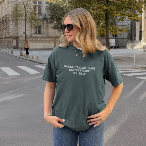 A woman wearing a dark green graphic t-shirt with the text 'HATING TAYLOR SWIFT DOESN'T MAKE YOU DEEP' stands on a city street in front of a historic building.