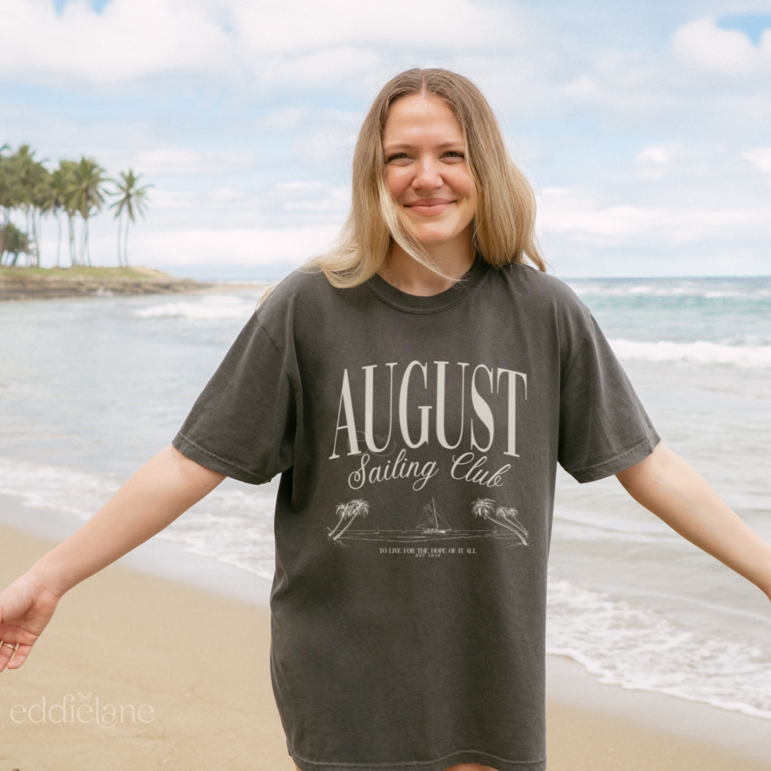 Woman wearing a minimalistic graphic tee that says 'August Sailing Club' with a sketch design of sailboats on a beach. The woman is standing on a beach with palm trees and othe cean in the background. Inspired by August from Folklore by Taylor Swift.