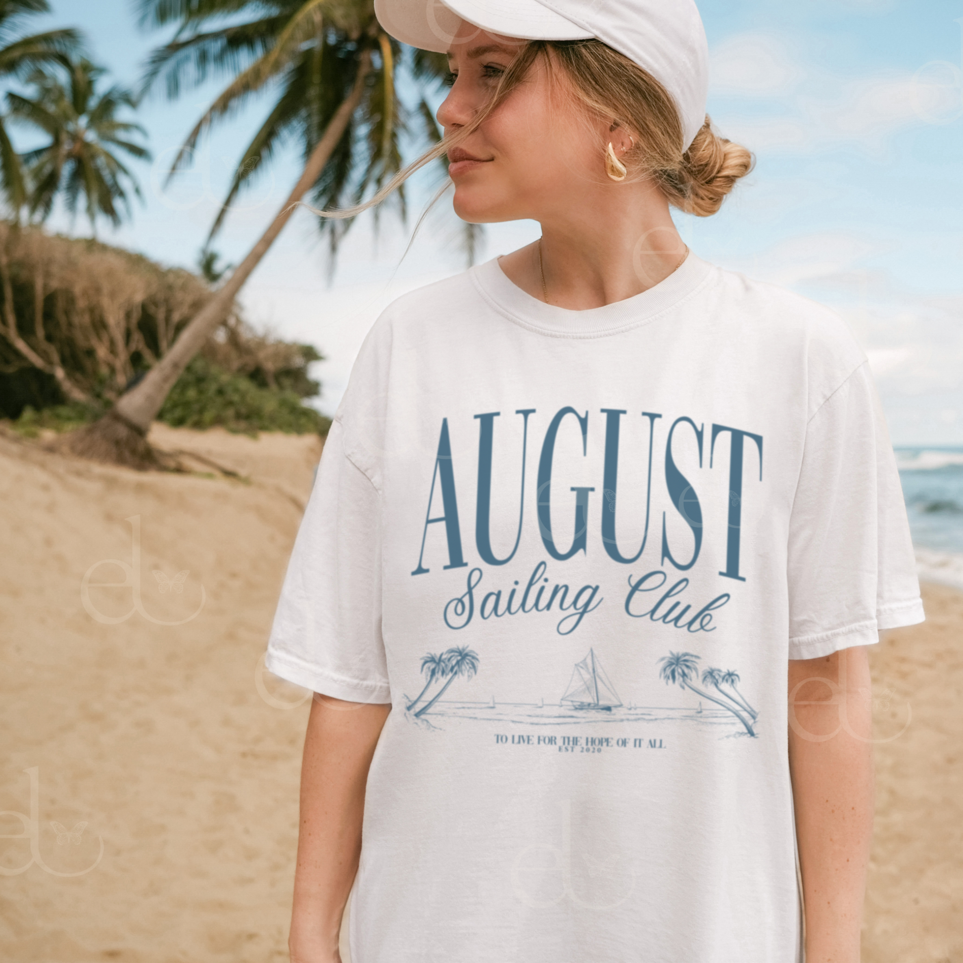Woman wearing a minimalistic graphic tee that says 'August Sailing Club' with a sketch design of sailboats on a beach. The woman is standing on a beach with palm trees and othe cean in the background. Inspired by August from Folklore by Taylor Swift.