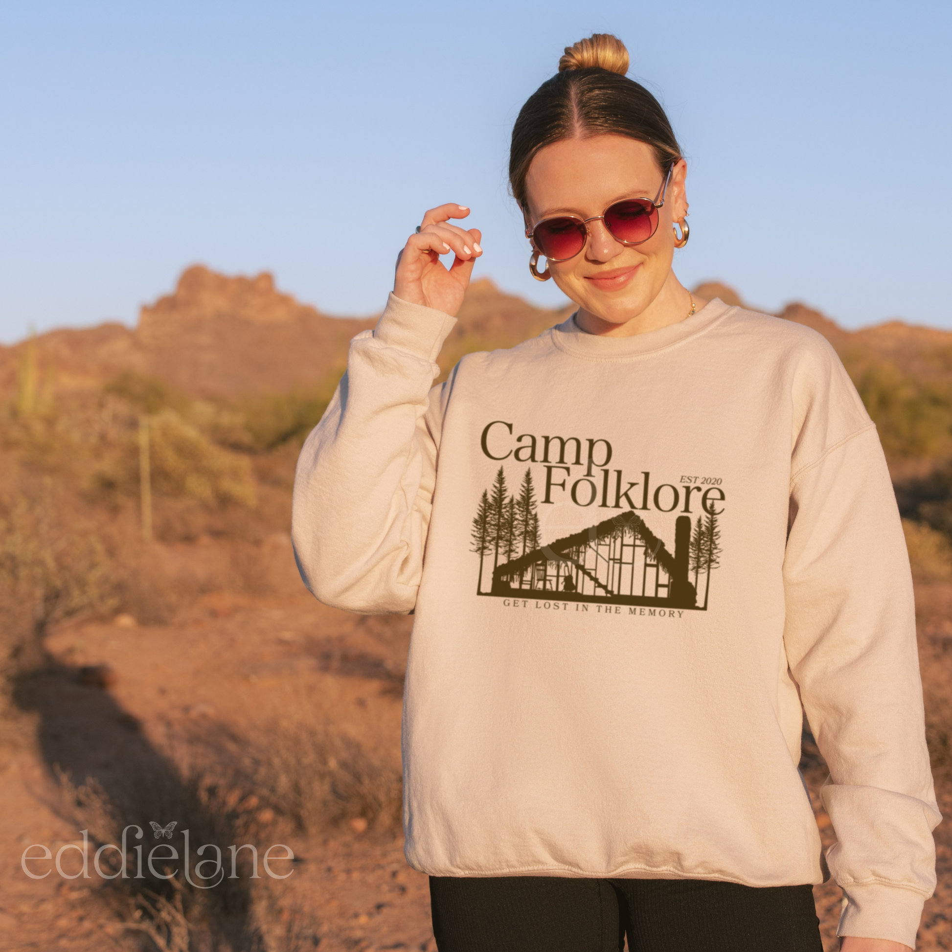 Woman wearing a sand-colored sweatshirt that says "Camp Folklore" with a silhouette of the Folklore cabin inspired by Taylor Swift's Folklore.