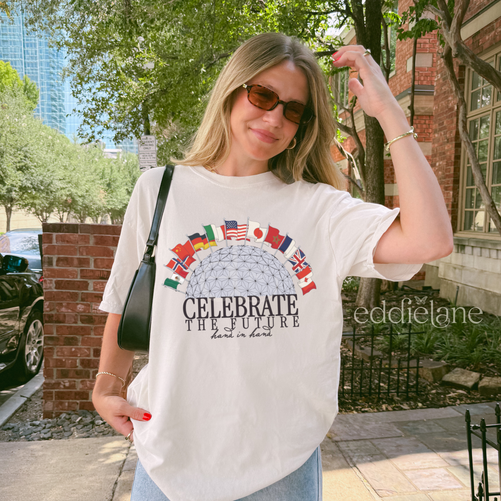 Woman wearing an ivory comfort colors t-shirt with a graphic design of an epcot ball surrounded by 12 country flags and the words "celebrate the future hand in hand"