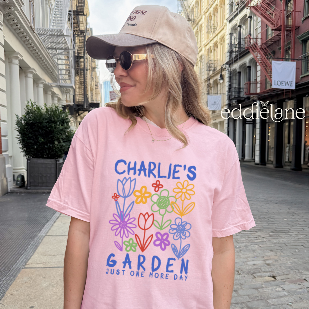 woman wearing a pink comfort colors shirt with a doodle design of flowers in different colors that says "charlie's garden just one more day"