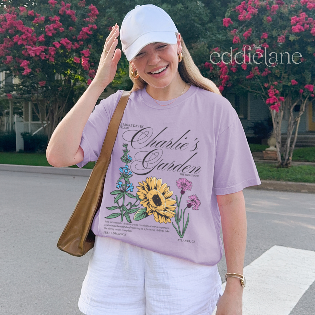 Person wearing a light purple graphic tee with a floral design that says 'Charlie's Garden' t-shirt with floral design,