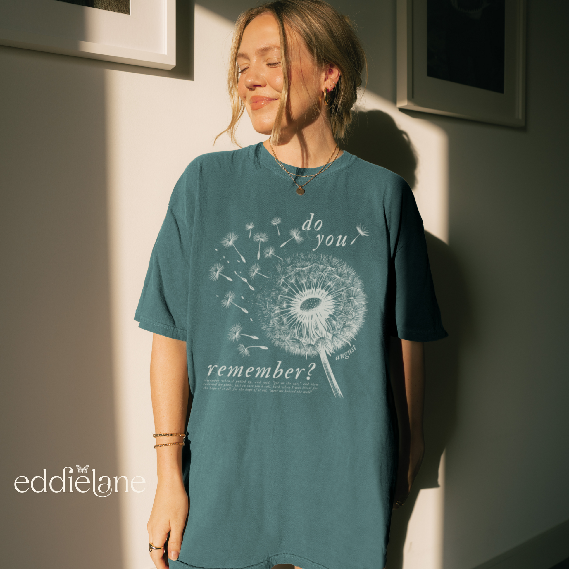 girl standing in the sunlight in a teal blue/green oversized t-shirt with a graphic design featuring a dandelion blowing in the wind, with the words "do you remember?". inspired by taylor swift folklore august.