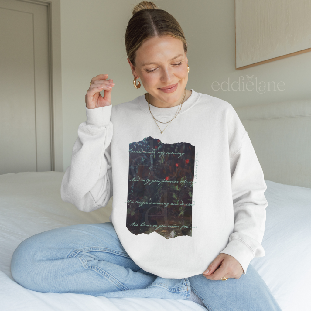 Woman wearing a white sweatshirt with a picutre of a painting and the words "Locked inside my memory and only you possess the key no longer drowning and deceived all because you came for me"