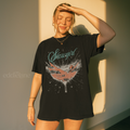woman wearing an oversized black t-shirt featuring a graphic of a woman in a champagne glass surrounded by sparkles