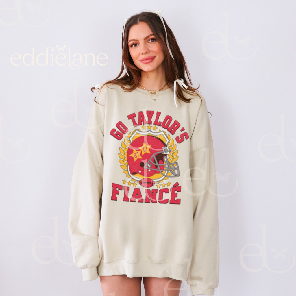 woman wearing an oversized sand crewneck sweatshirt with a vintage football helmet design and text that says "go taylor's fiance"