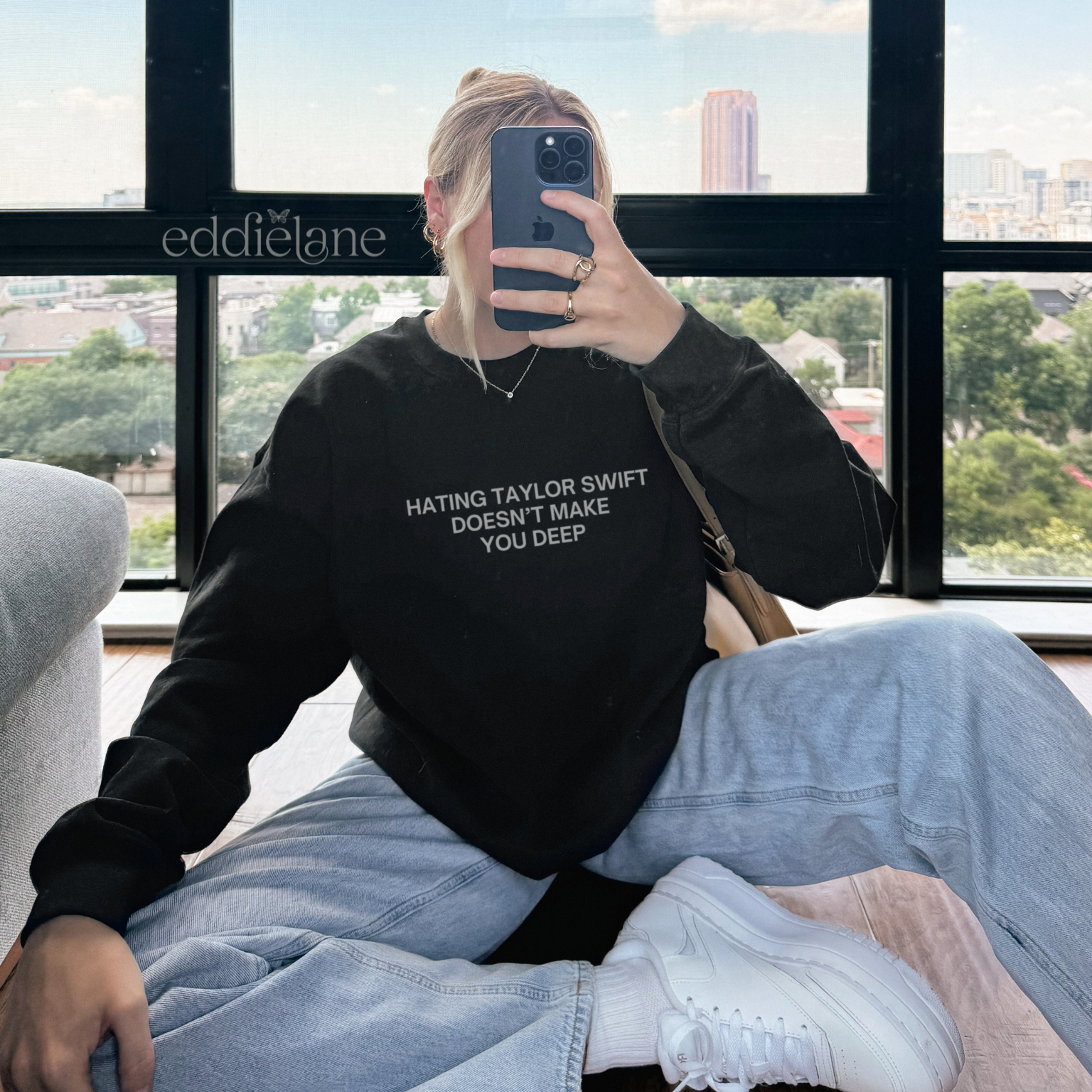 Woman wearing a black sweatshirt with text that says "hating taylor doesn't make you deep", sitting in front of a large window with a cityscape view.