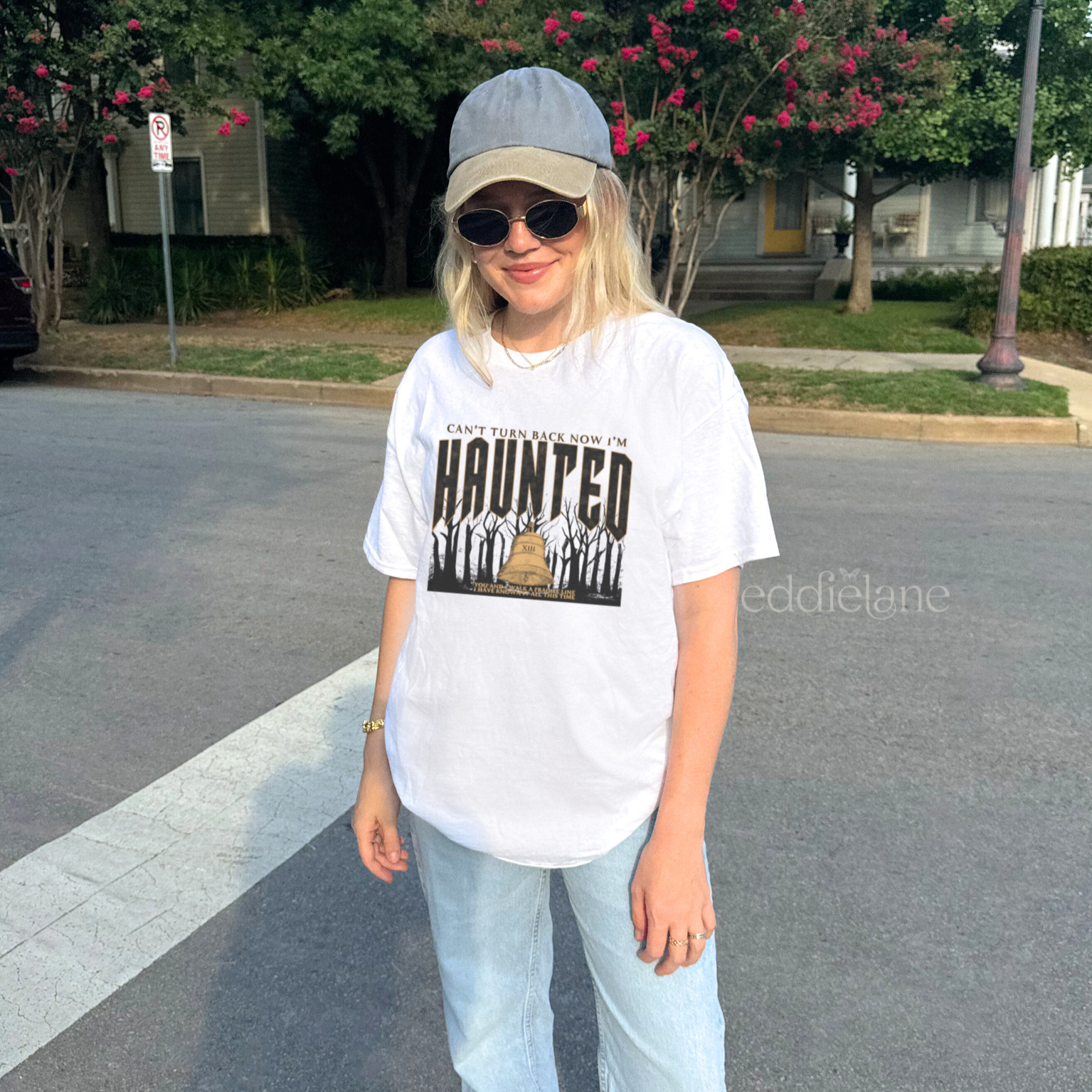 woman wearing a white graphic tee that says "can't turn back now i'm haunted" with a graphic of spooky trees and a bell