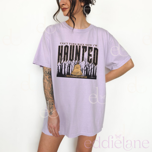 woman wearing an oversized light purple graphic tee that says "can't turn back now i'm haunted" with a graphic of spooky trees and a bell