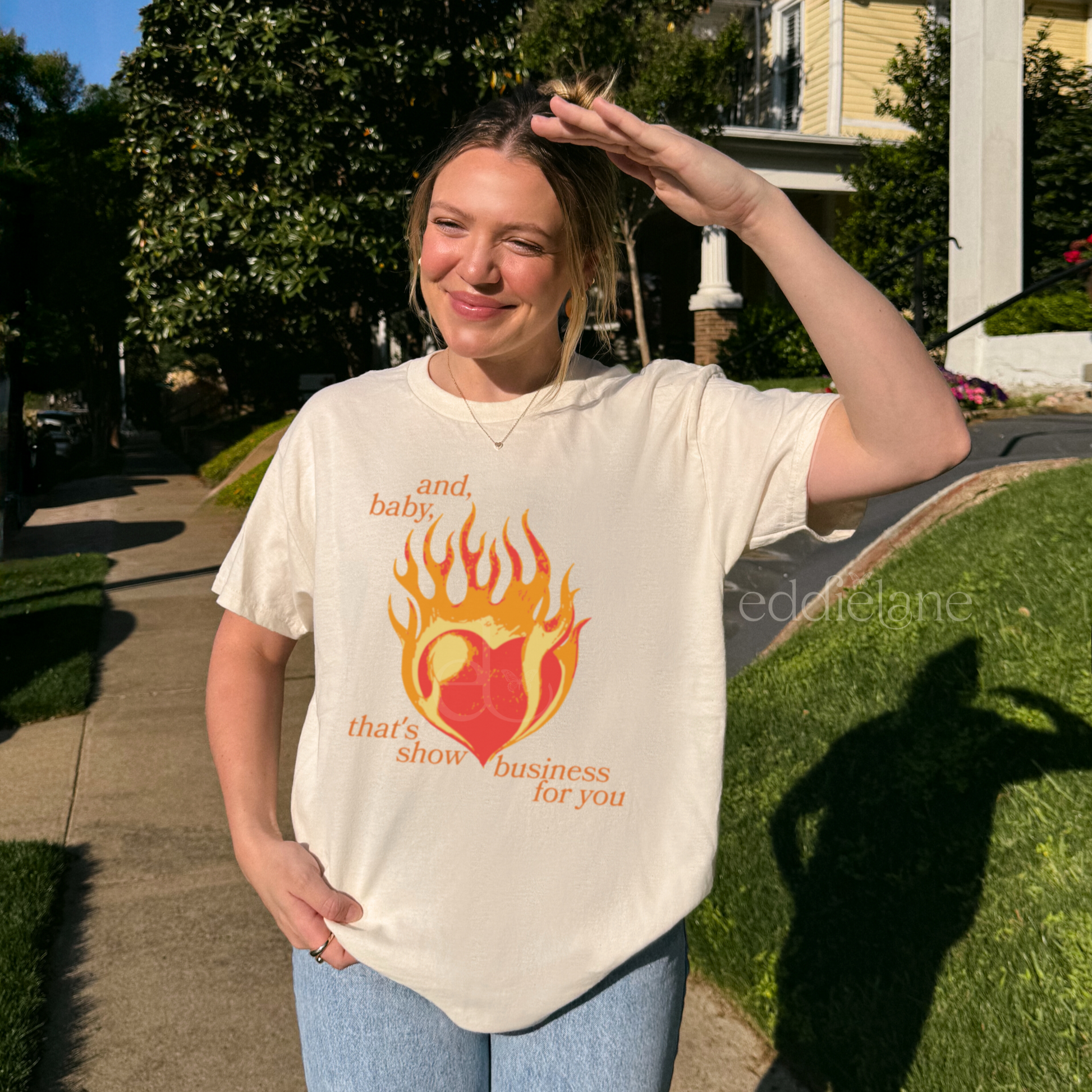 Woman wearing a cream t-shirt with a heart in flames graphic and the words "and, baby, that's show business for you" inspired by taylor swift.
