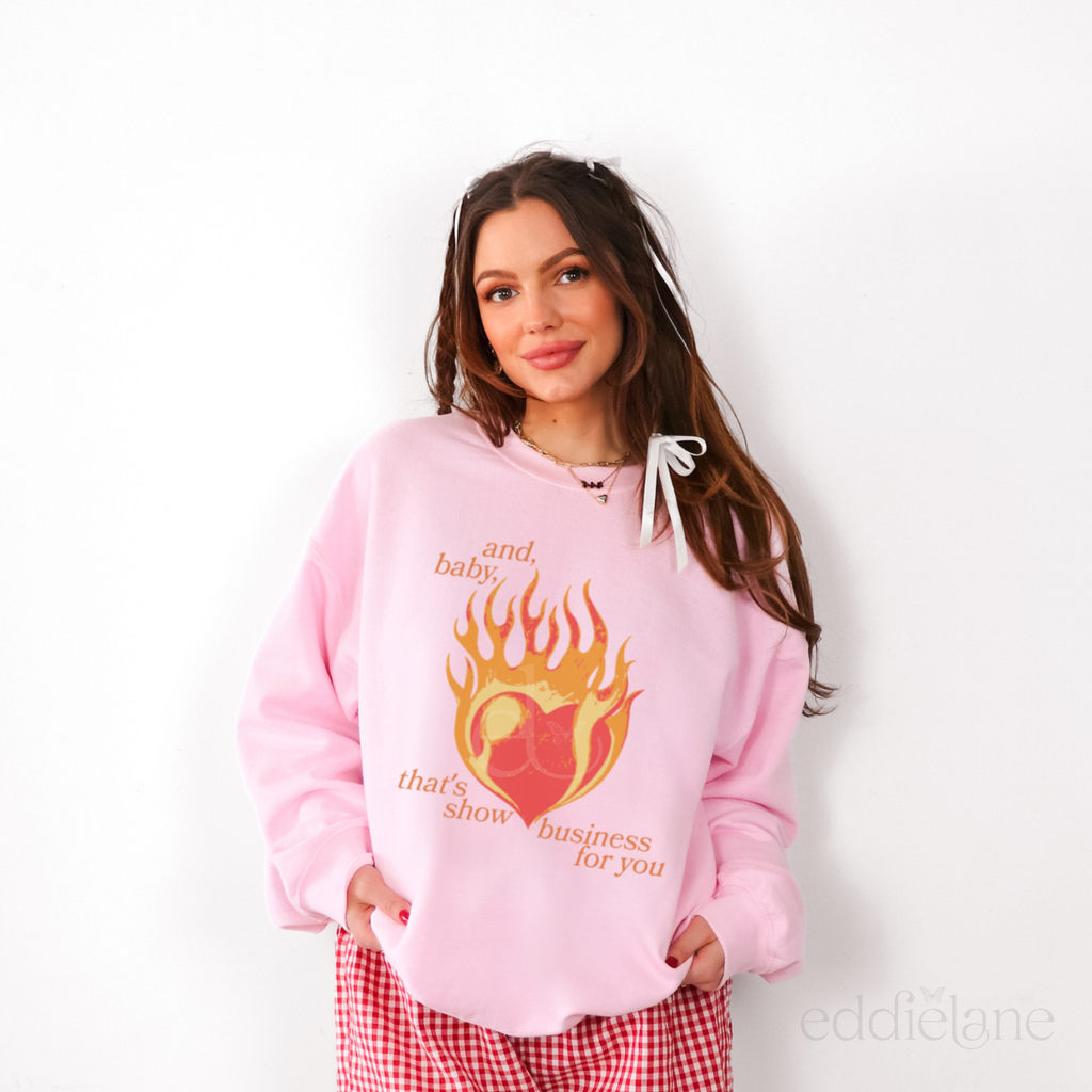 Woman wearing a pink hoodie with a graphic design of a heart on fire and the words "and baby thats show business for you"