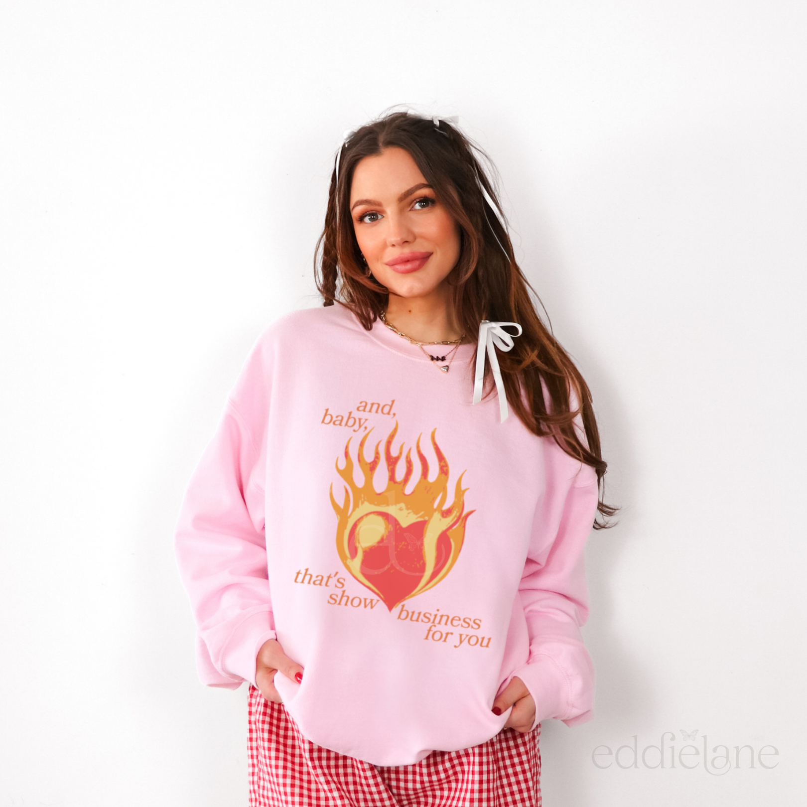 Woman wearing a pink hoodie with a graphic design of a heart on fire and the words "and baby thats show business for you"