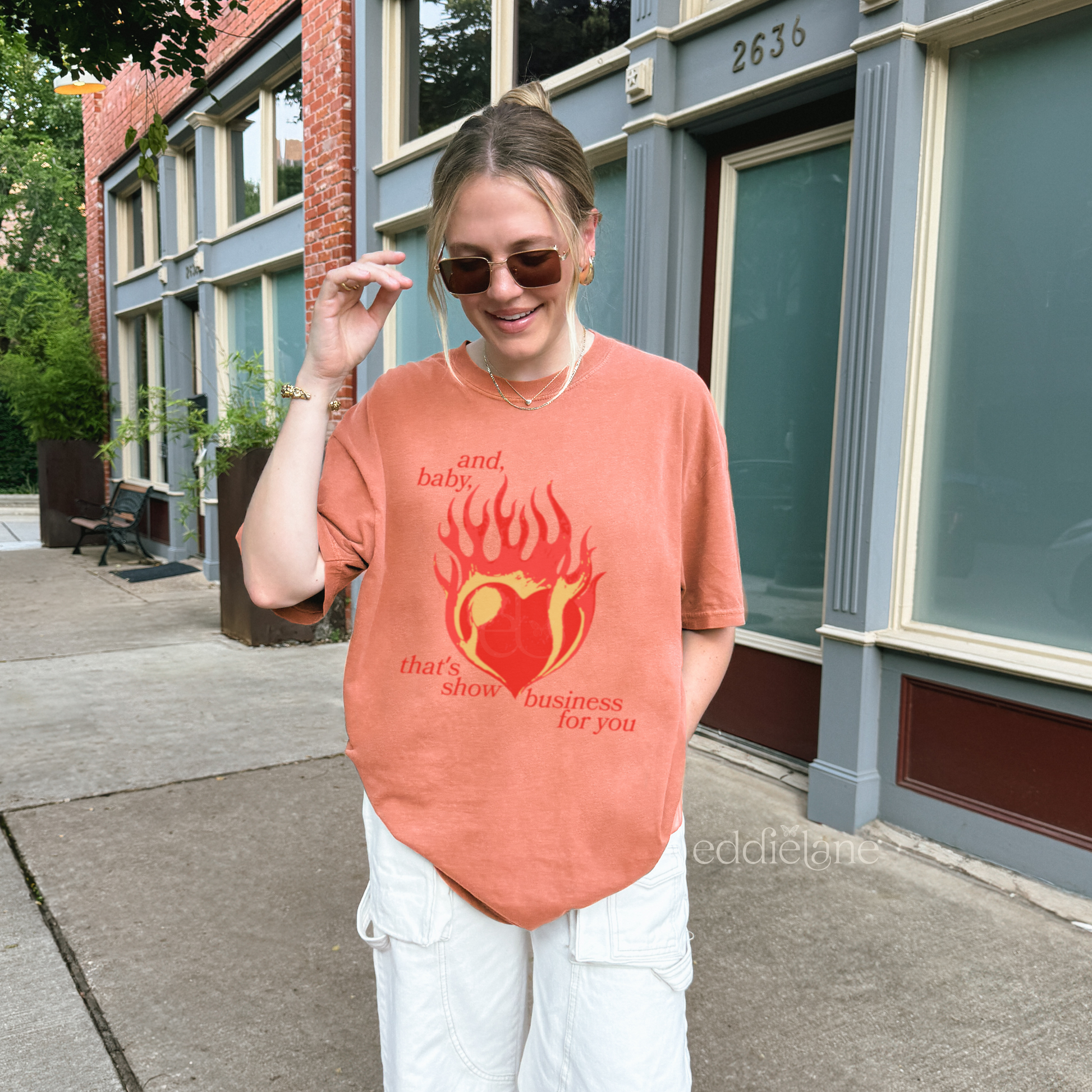 Woman wearing a light orange t-shirt with a heart in flames graphic and the words "and, baby, that's show business for you" inspired by taylor swift.