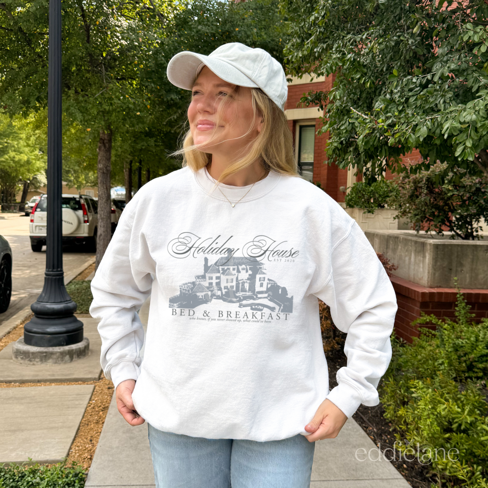 Woman wearing a white crewneck sweatshirt that says "Holiday House Bed and Breakfast" with a silhouette of the holiday house from taylor swift's the last great american dynasty from folklore.
