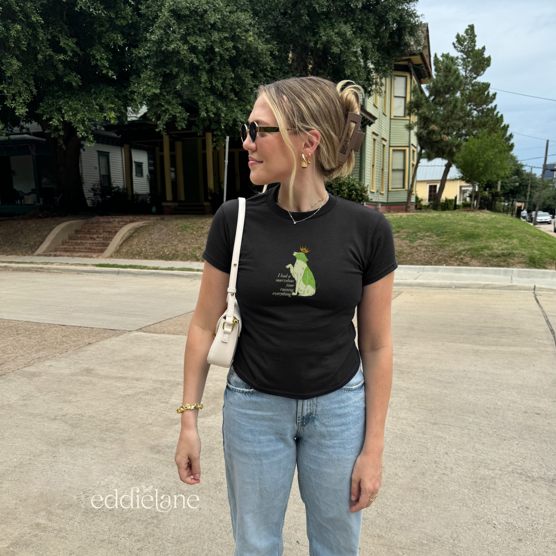 Girl wearing a black baby tee with a graphic of a key-lime green dog wearing a crown and the words "i had a marvelous time ruining everything", standing outdoors. Inspired by the song the last great american dynatsy from folklore by taylor swift.