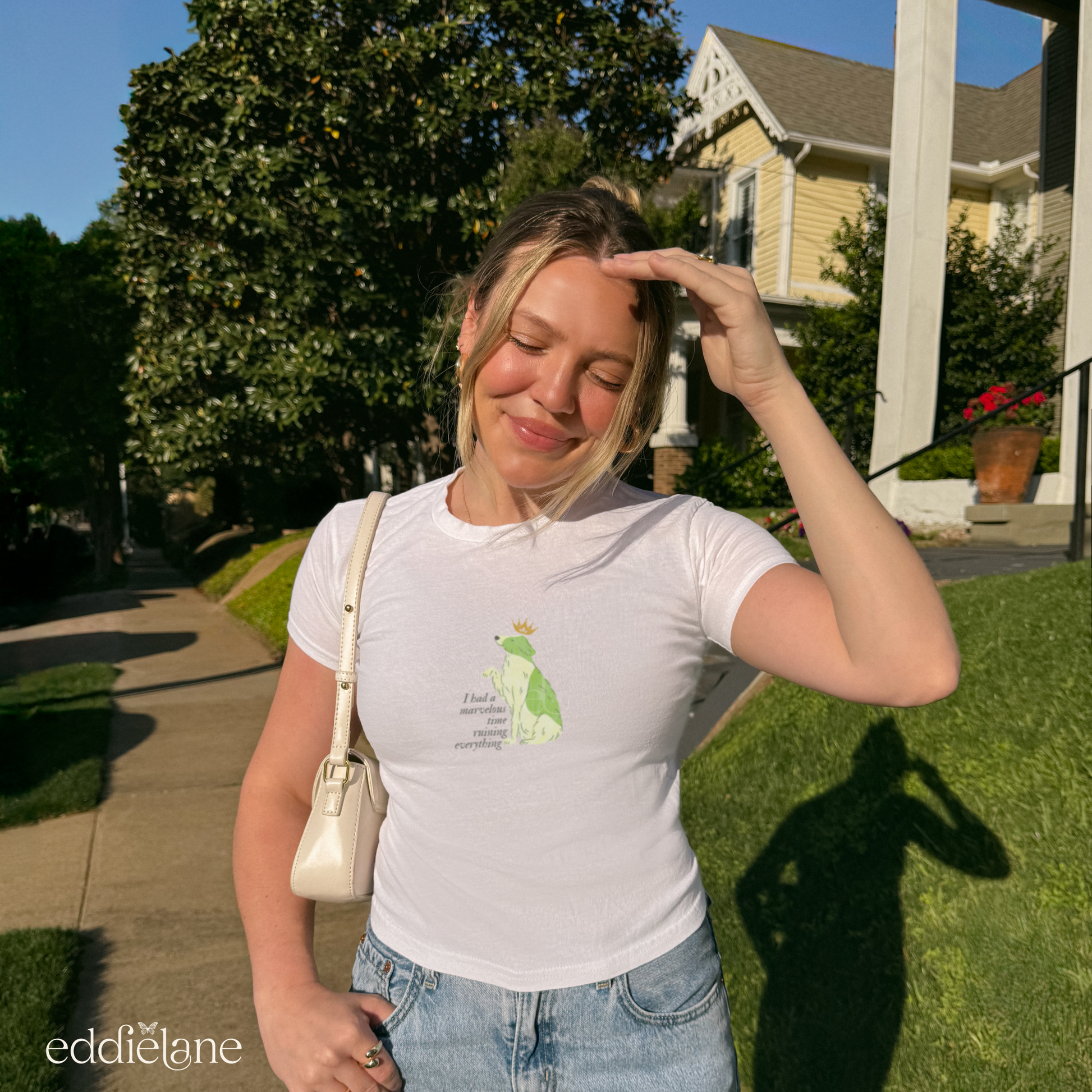 Girl wearing a white baby tee with a graphic of a key-lime green dog wearing a crown and the words "i had a marvelous time ruining everything", standing outdoors. Inspired by the song the last great american dynatsy from folklore by taylor swift.