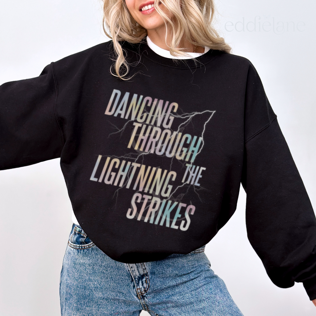 Woman wearing a black sweatshirt with the words "dancing through the lightning strikes" and a colorful lightning bolt design
