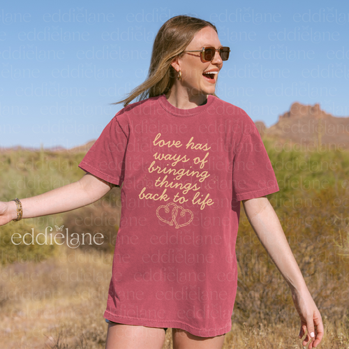 Woman wearing a red t-shirt with text and a heart design, standing in a desert landscape.