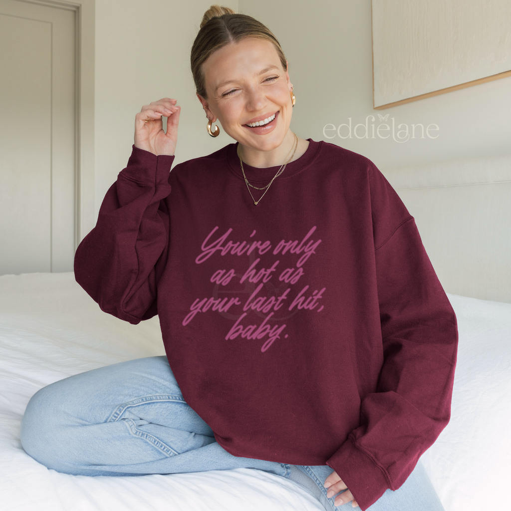 woman in a maroon crewneck sweatshirt that says "you're only as hot as your last hit, baby" in pink.