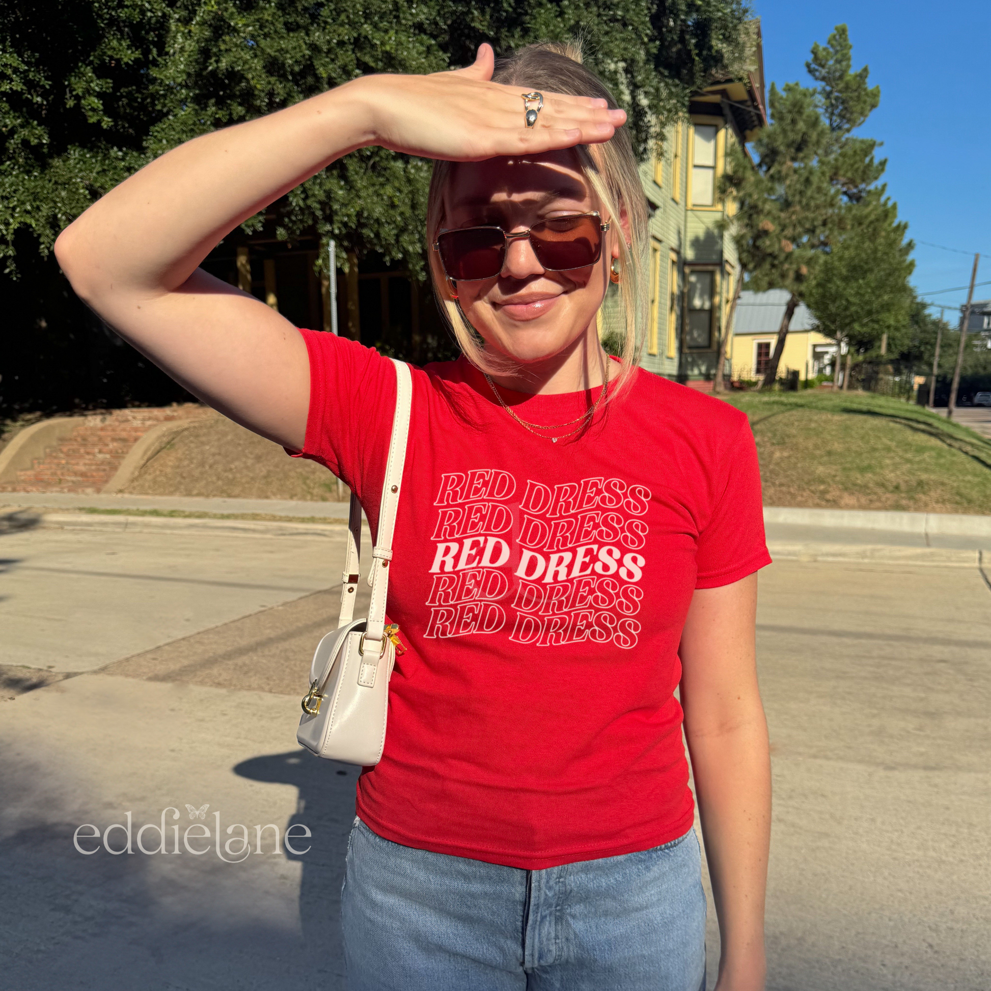 Woman wearing a red baby tee with "red dress" written in a curvy design inspired by the jonas brothers song burnin up.