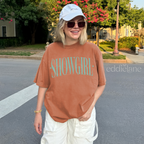 Woman wearing an orange comfort colors graphic tee that says 'SHOWGIRL' with two small stars