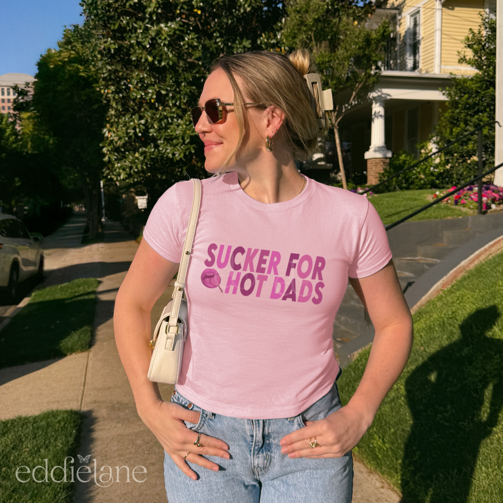 Woman wearing a pink baby tee with dark pink design that says "sucker for hot dads" 