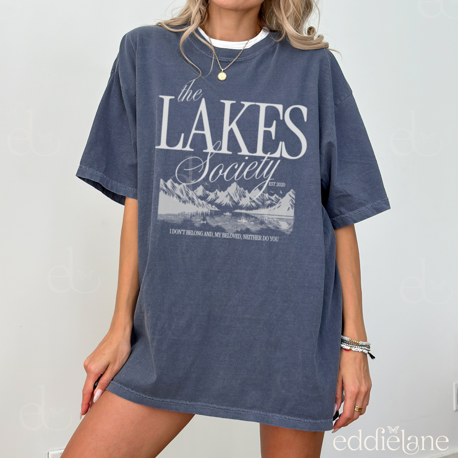 Girl wearing an oversized blue graphic t-shirt that says 'The Lakes Society' wth a sketch of a lake surrounded by mountains and the words "i don't belong and my beloved neither do you" inspired by folklore, taylor swift, and the lakes.