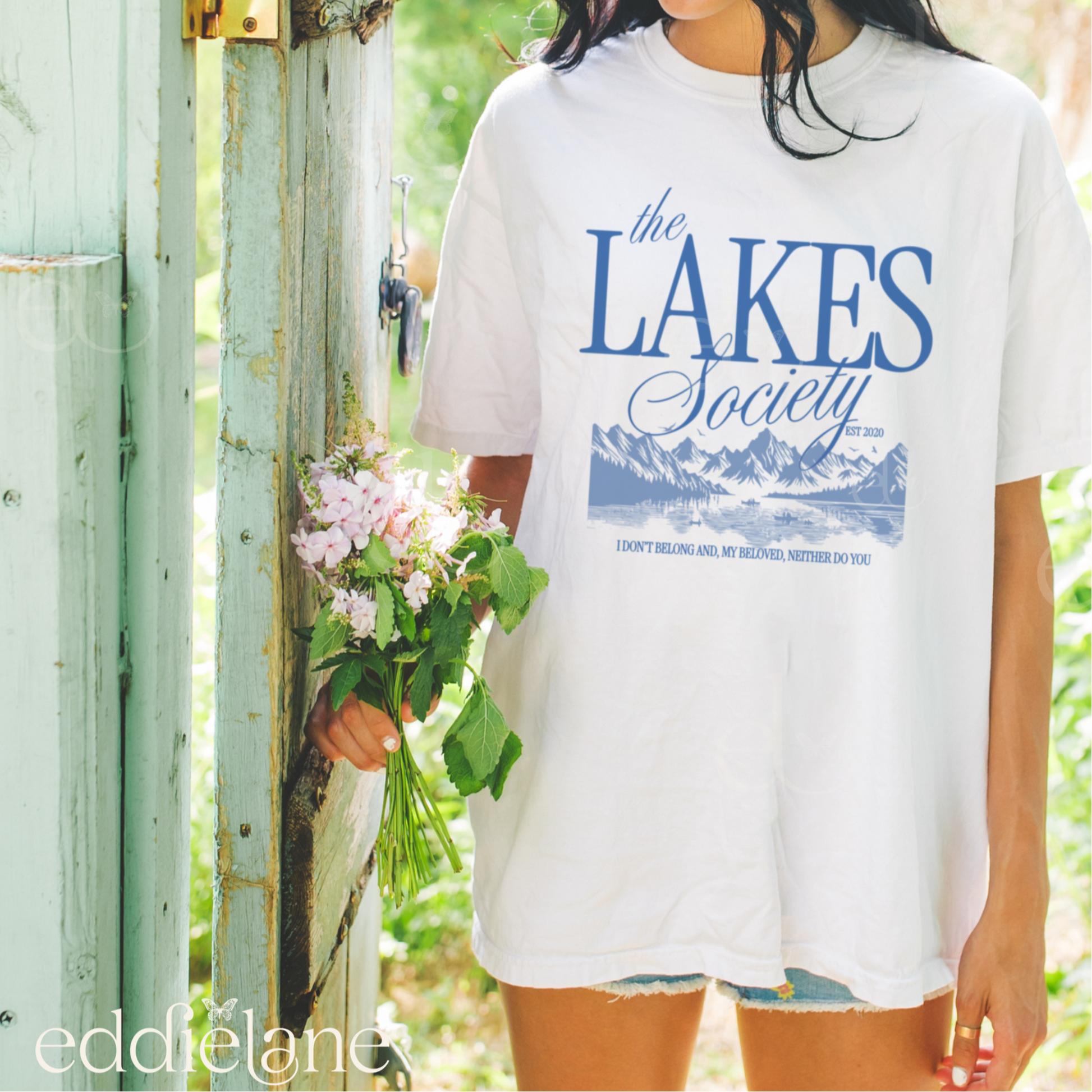 Girl wearing an oversized white graphic t-shirt that says 'The Lakes Society' wth a sketch of a lake surrounded by mountains and the words "i don't belong and my beloved neither do you" inspired by folklore, taylor swift, and the lakes.