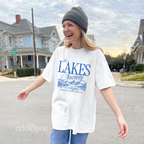 Girl wearing an oversized white graphic t-shirt that says 'The Lakes Society' wth a sketch of a lake surrounded by mountains 