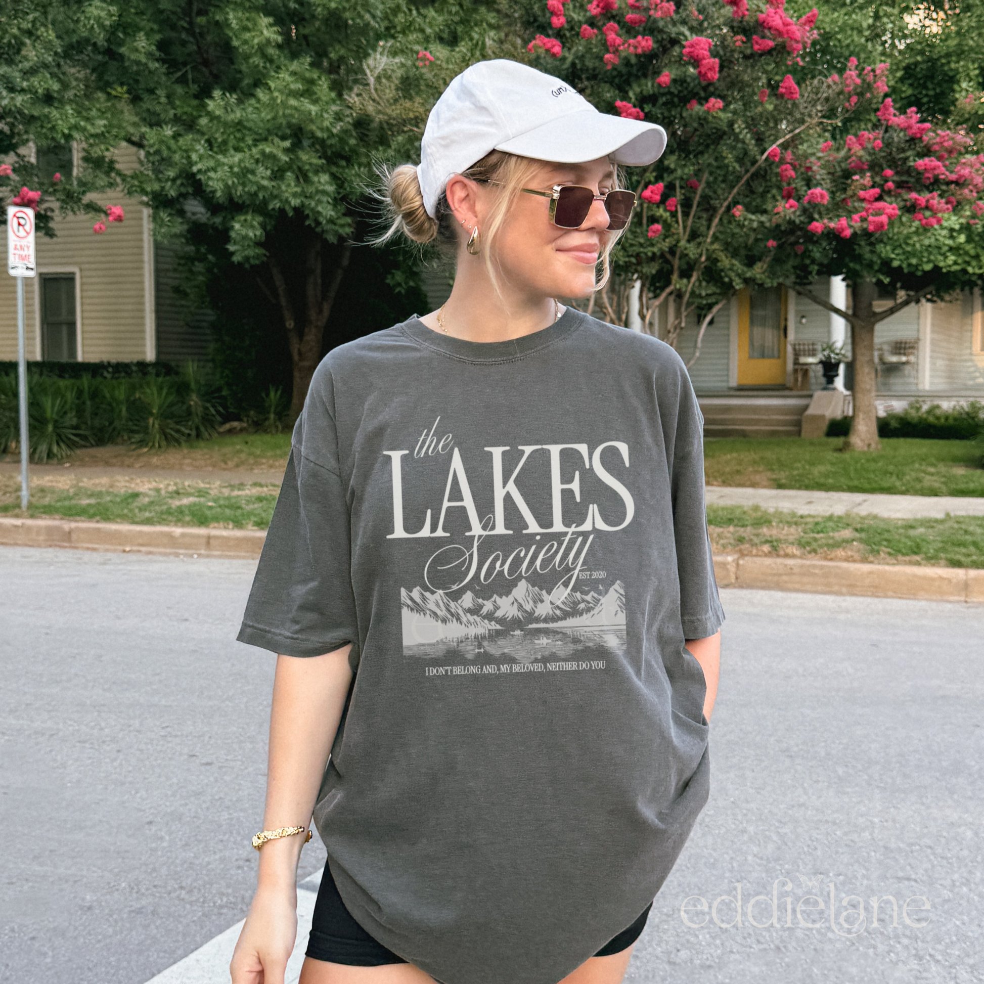 Girl wearing an oversized gray graphic t-shirt that says 'The Lakes Society' wth a sketch of a lake surrounded by mountains and the words "i don't belong and my beloved neither do you" inspired by folklore, taylor swift, and the lakes.