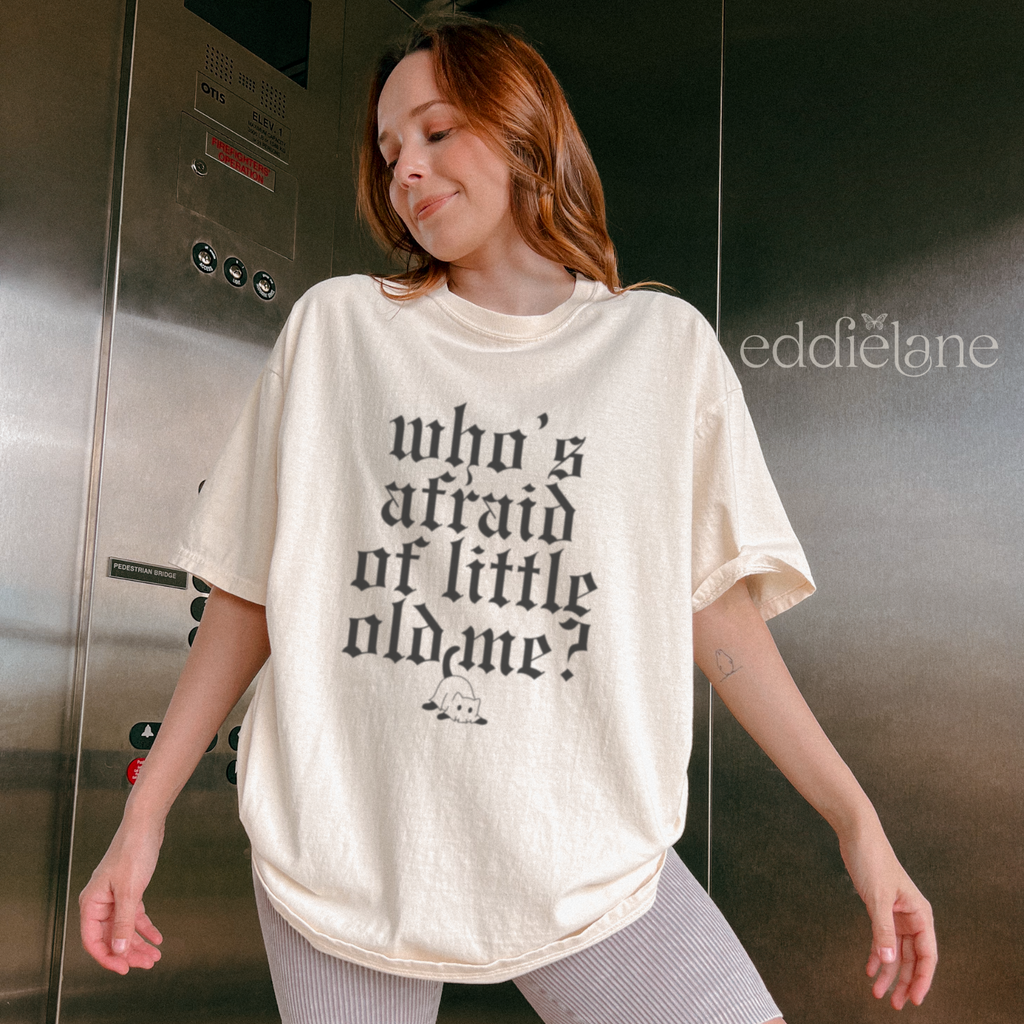 Woman wearing an ivory comfort colors t-shirt that says "who's afraid of little old me" with a graphic of a cat wearing a ghost costume