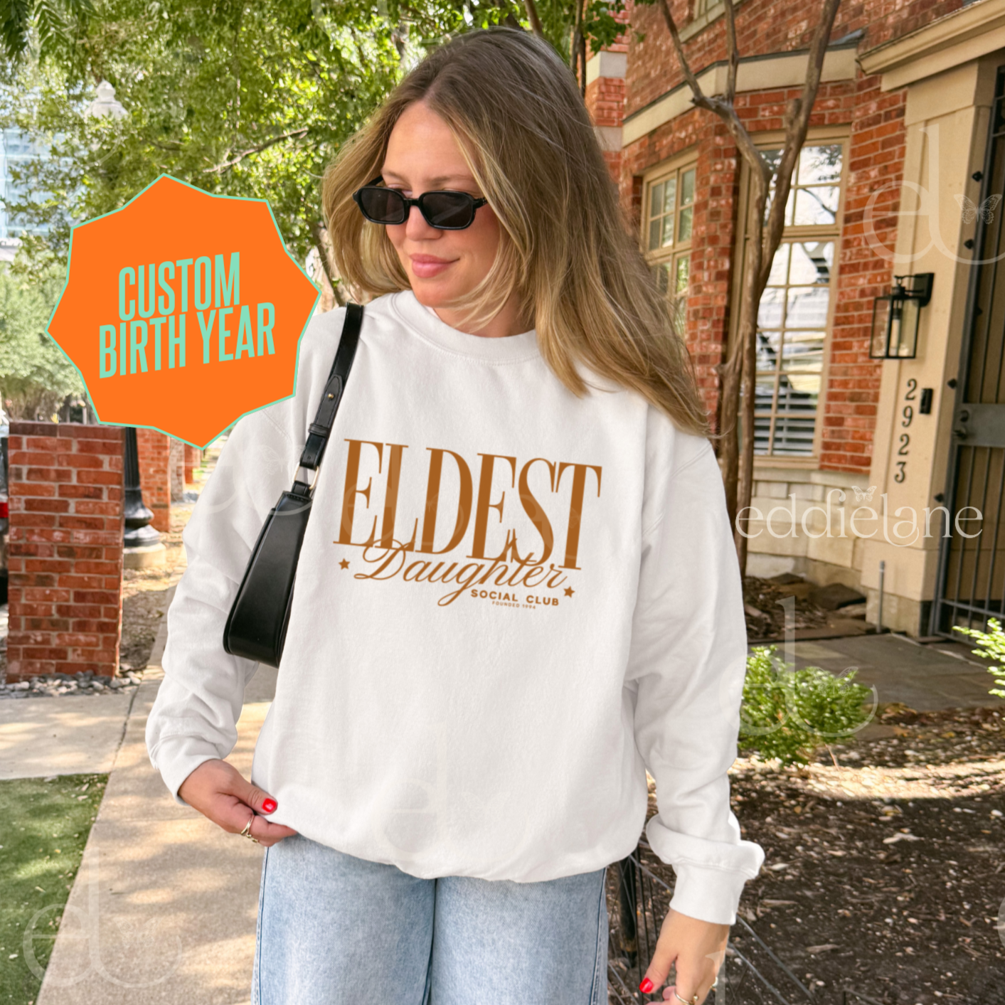 Woman wearing a white sweatshirt that says 'Eldest Daughter social club'.