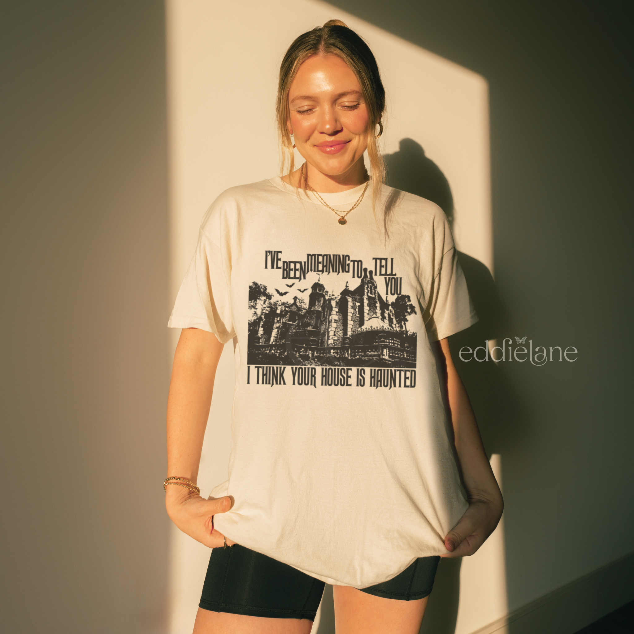 woman wearing an oversized ivory comfort colors halloween t-shirt with a graphic of haunted mansion and the words "i've been meaning to tell you I think your house is haunted"